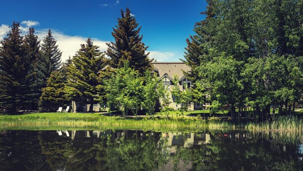 A serene lakeside view with a row of traditional-style buildings, a clear sky, and lush greenery. - 3BR Pet Friendly - Ski Hill Rd (Driggs)