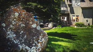 A house with a large rock in the foreground, a well-maintained lawn, and a deck area with outdoor furniture. - 3BR Pet Friendly - Ski Hill Rd (Driggs)