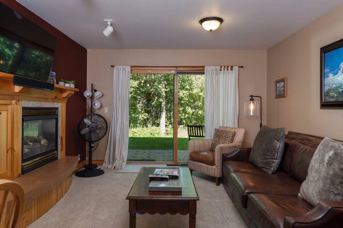 A living room with a fireplace, a sofa, a coffee table, and a view of a green garden through a sliding glass door.