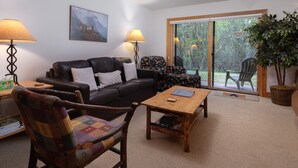 Un salon comprenant un canapé, une table basse en bois, un fauteuil à motifs et une grande fenêtre donnant sur un paysage verdoyant.