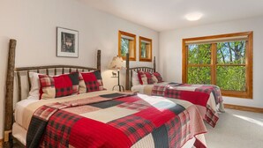 A room with two beds, a wooden headboard, a lamp, a framed picture, and a window with a view of trees.