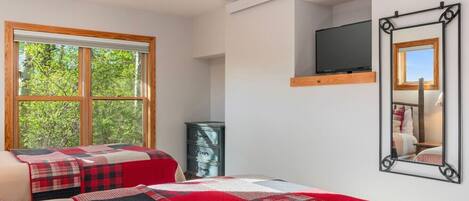 Un dormitorio con un ventanal grande, una cama con colcha roja y negra, un aparador y un televisor montado en la pared.