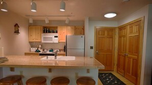 A kitchen with wooden cabinets, a white microwave, a refrigerator, a sink, and three wooden stools. - Grizzly Bear Den: Near Targhee, Sleeps 6, Hot Tub (Driggs)
