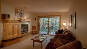 Un salon chaleureux avec une cheminée, un canapé, une table basse et une grande fenêtre donnant sur les arbres.