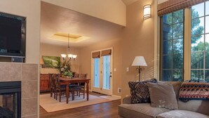 A spacious living room with a fireplace, a dining area with a wooden table and chairs, and a large window with a view of trees. - Alpine Meadows: Creekside, Aspen Views, Light & Spacious, Fireplace & 2-car garage (Driggs)