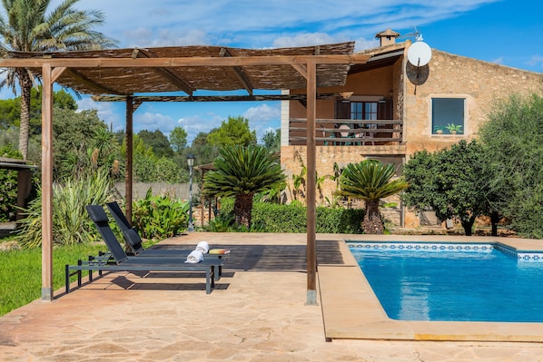Un espace aménagé au bord de la piscine, avec des chaises longues, une pergola et un bâtiment en pierre doté d’un balcon.