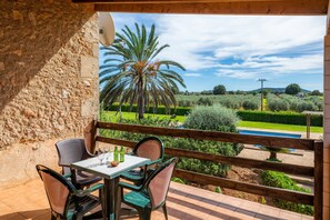 A balcony with a table and chairs, overlooking a landscaped garden with a palm tree and a pool. - Vacation Home 'Cas Casai' with Mountain View, Pool & Wi-Fi (Son Negre)
