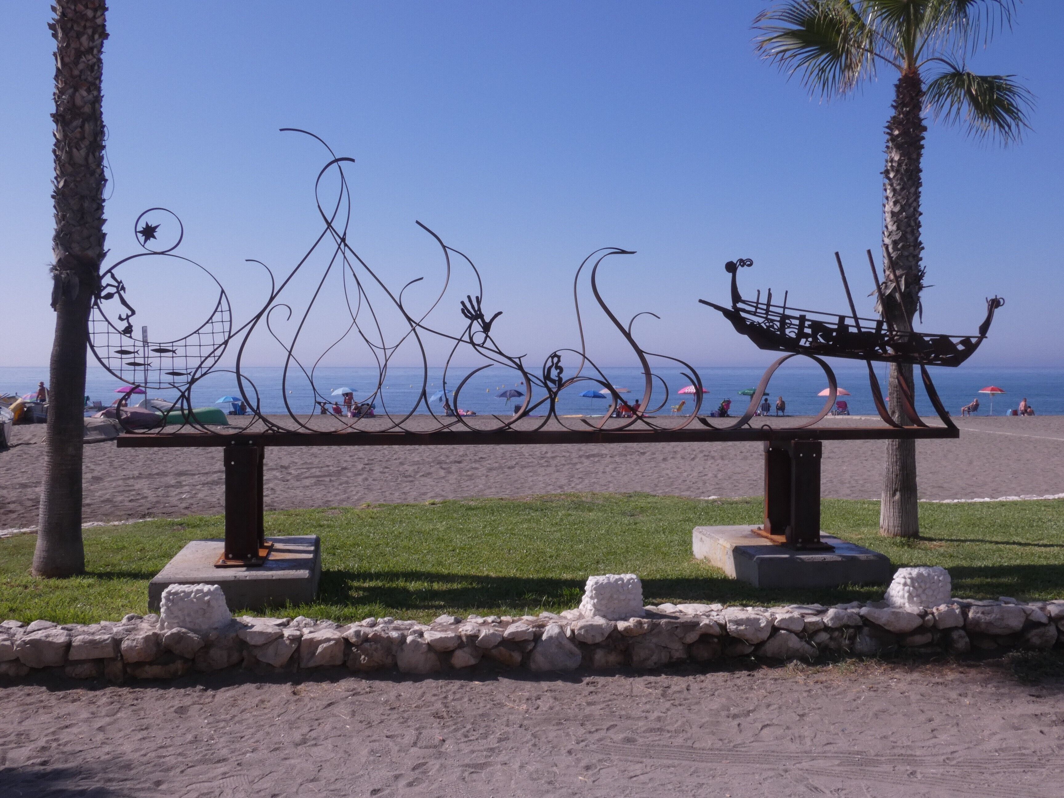 A beach bench with a decorative metal sculpture, surrounded by palm trees and a clear view of the ocean.