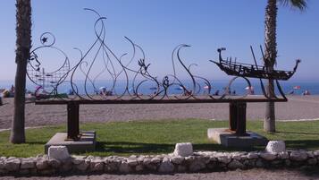 A beach bench with a decorative metal sculpture, surrounded by palm trees and a clear view of the ocean.
