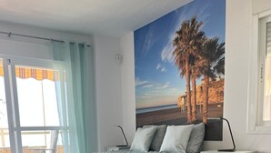 Un dormitorio con una cama, una mesita de noche, una pequeña mesa y vistas a la playa a través de una ventana.