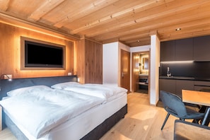 A modern bedroom with a large bed, a TV mounted on the wall, and a separate kitchen area.