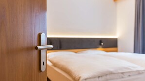Habitación de hotel con puerta de madera, cama con ropa blanca, cabecero y mesita de noche.