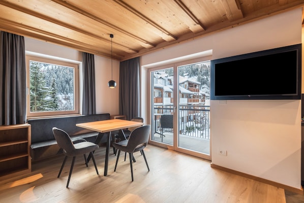 A modern interior with a wooden ceiling, a dining table with chairs, a TV mounted on the wall, and large windows with a view of a snowy landscape.