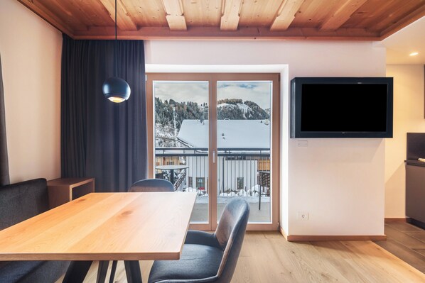 A modern interior with a wooden ceiling, a dining table with chairs, a large TV mounted on the wall, and a sliding glass door leading to a balcony with a snowy view.