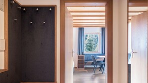 A hallway with a wooden bench, a dark wall with hooks, and a view into a room with a desk and chair.