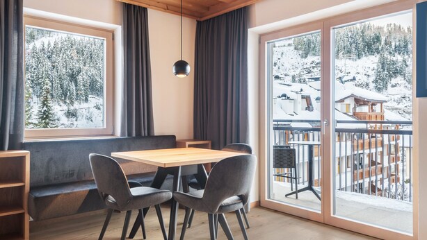 A room with a wooden ceiling, a wooden table with four chairs, a bench, and large windows with a snowy landscape view.