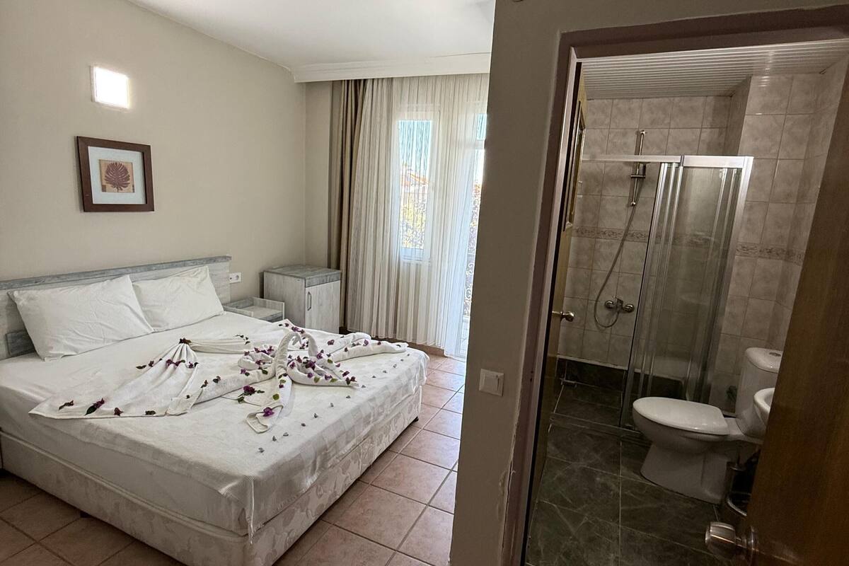 A bedroom with a bed, a bedside table, a nightstand, a framed picture on the wall, and a bathroom with a shower, toilet, and glass-enclosed shower area.