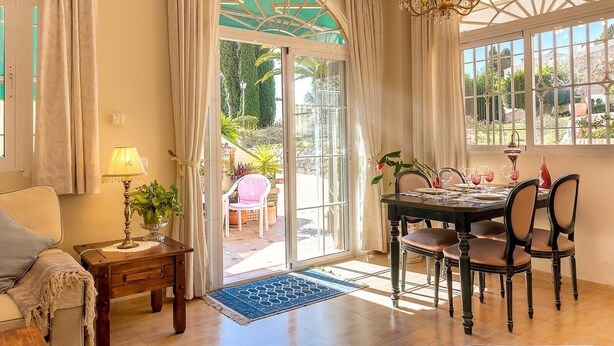 A dining area with a table set for four, a chandelier, and a view of the garden through large windows. - Apartment "Capricho Montepino 1 Las Palomas" with Pool, Wi-Fi, A/C, Terraces & Garden (La Herradura)