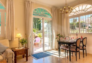 A dining area with a table set for four, a chandelier, and a view of the garden through large windows.