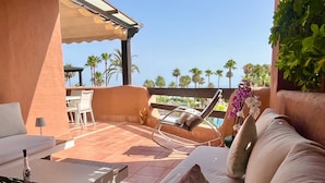 A sunlit outdoor seating area with a cushioned sofa, a metal chair, and a table with a bottle and a plant. - Beach front apartment with Sea View (Estepona)