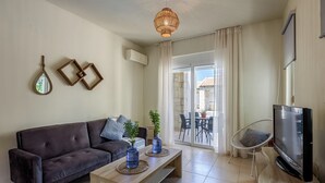 A modern living room with a dark brown sofa, wooden coffee table, and a television on a wooden stand. There are two blue vases on the coffee table, and a wicker chair is visible in the background. - AZURE VILLA -BEACH FRONT  (XANIA)