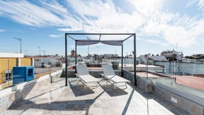 Terraza en la azotea con dos sillones de descanso bajo una pérgola, barandilla de cristal y vista panorámica de la ciudad.