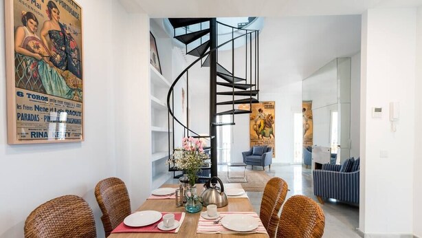 Un comedor con una mesa de madera dispuesta para cuatro personas, rodeada de sillas de mimbre. Una escalera en espiral negra conduce a un nivel superior.