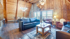 A spacious living area with a wooden ceiling, a black leather sofa, a wooden coffee table, and a large window providing natural light. - Lakefront A-Frame Cabin with Community Perks (Columbia)