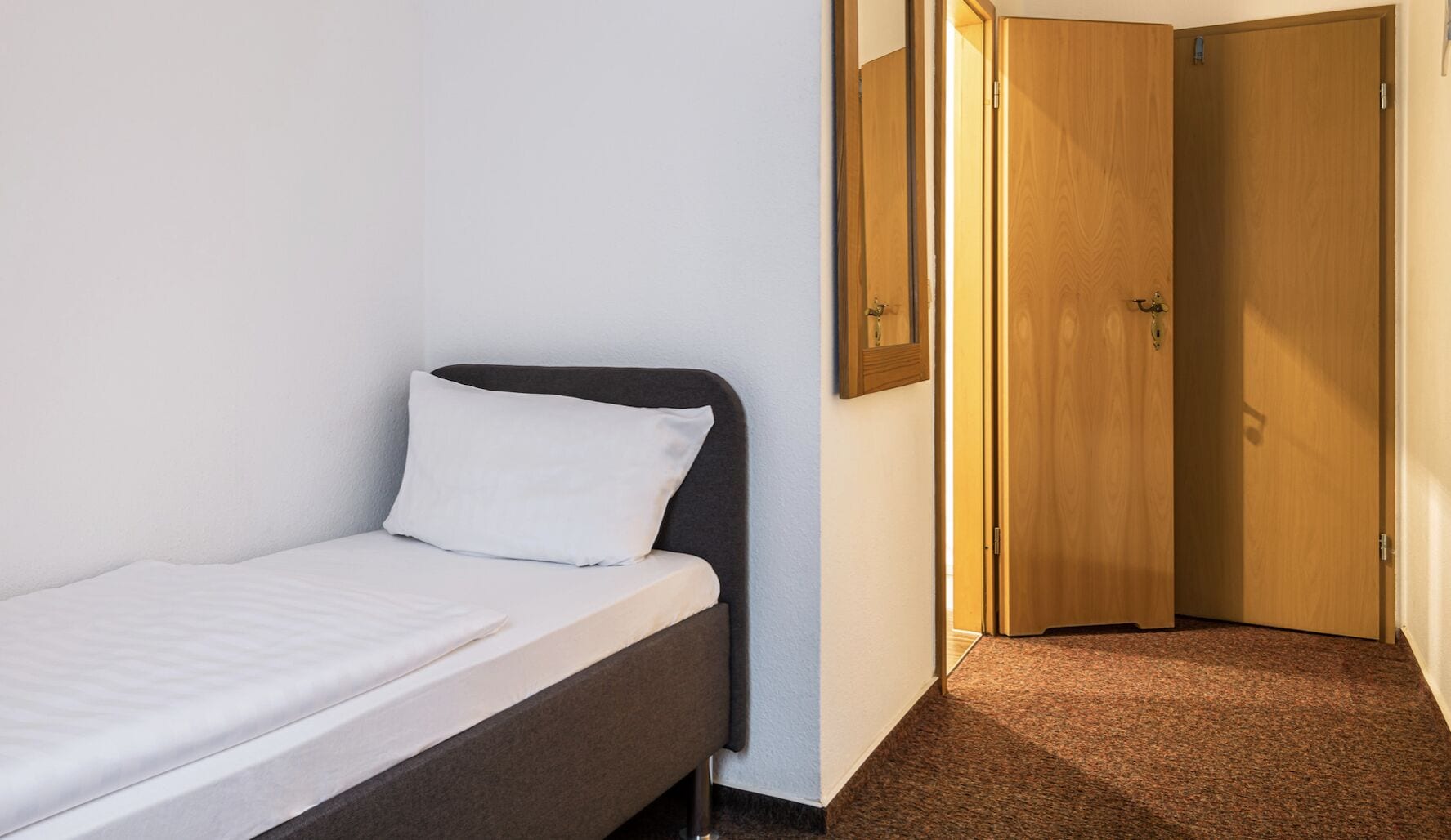 A small, single-bed room with a dark headboard, white bedding, a wooden door, and a carpeted floor.
