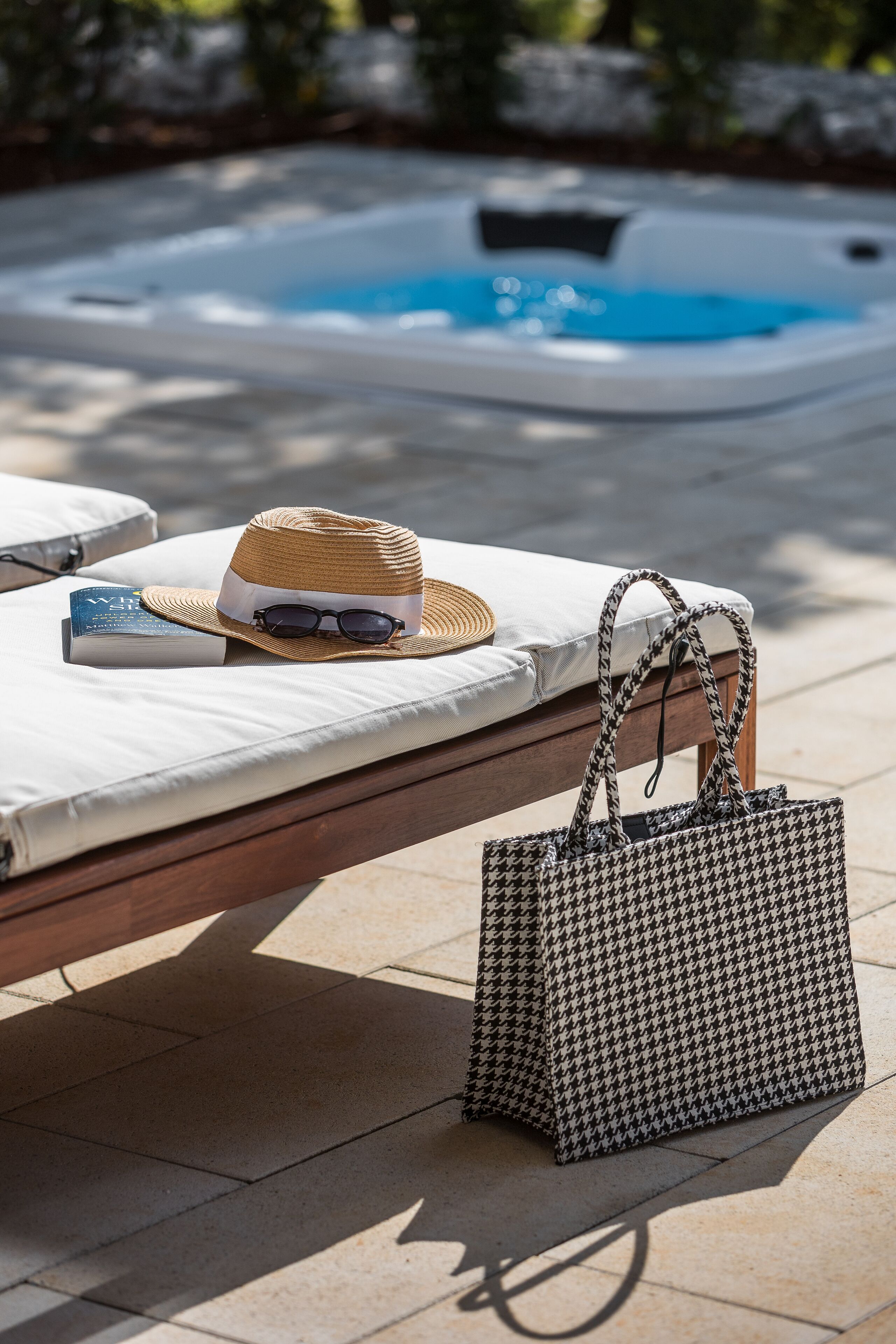 A sunbed with a hat, sunglasses, and a book, a patterned handbag, and a hot tub in the background.