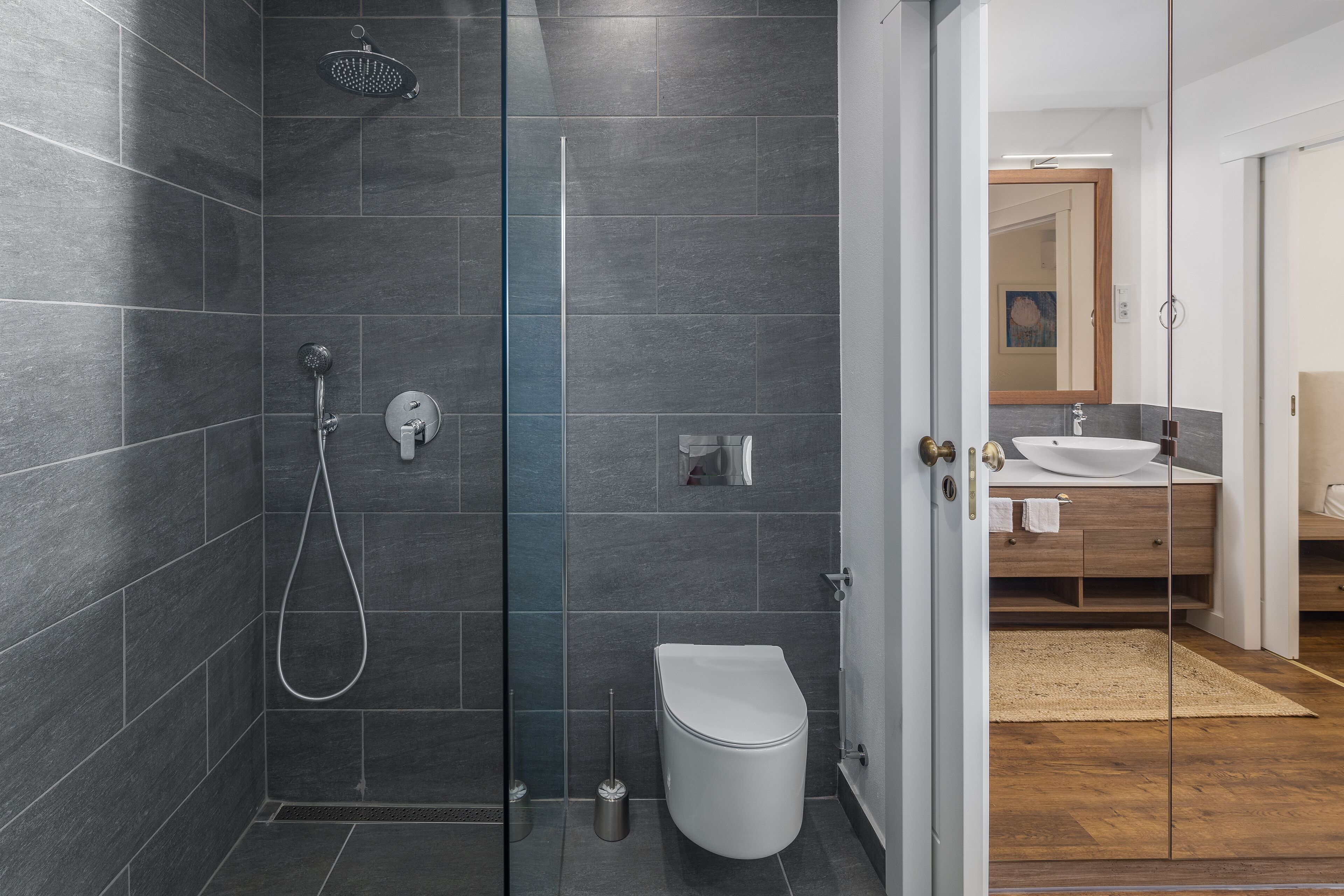 A modern bathroom with dark tiles, a glass shower enclosure, a white toilet, and a wooden vanity with a sink.