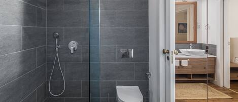 A modern bathroom with dark tiles, a glass shower enclosure, a white toilet, and a wooden vanity with a sink.