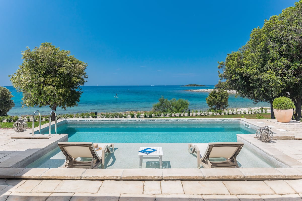 A swimming pool with lounge chairs and a table, overlooking a beach and the sea.