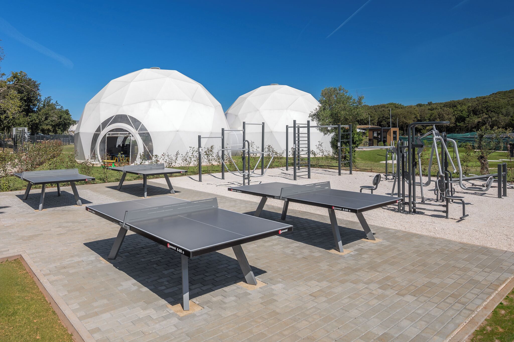 Outdoor exercise equipment, including tables and a climbing wall, in a park with white domed structures in the background.