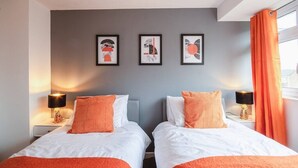 Camera d'albergo con due letti, entrambi con biancheria bianca e dettagli arancioni, una parete grigia con tre opere d'arte incorniciate e una finestra con tende arancioni.