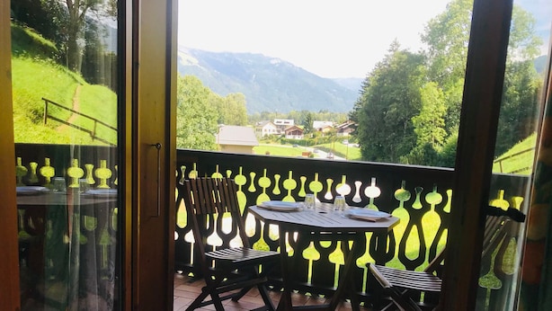 Un balcone con tavolo e sedie, una porta vetrata e vista su un paesaggio verde con montagne.