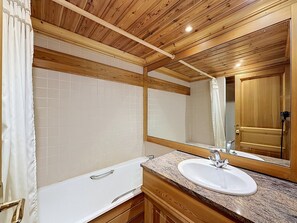 A bathroom with a wooden ceiling, white tiles, a white bathtub, a granite countertop, and a mirror.