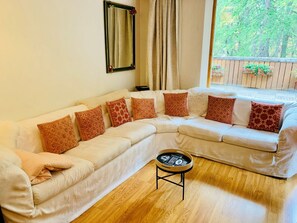 A L-shaped living room with a white sofa, patterned cushions, a small coffee table, a mirror, and a window with a view of greenery. - Charming 4-room apartment for 6 at slopes' edge with wifi (Val-d'Isère)