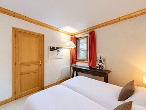 A bedroom with a double bed, a wooden door, a window with curtains, a bedside table, and a wall-mounted lamp.
