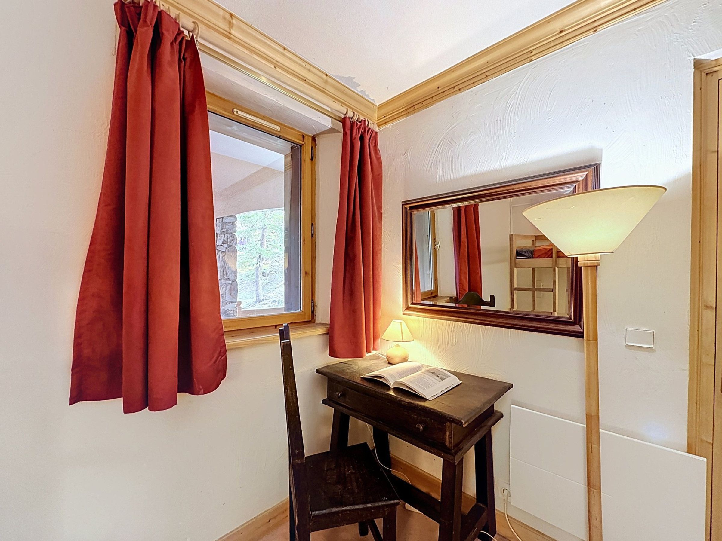 A room with a desk, chair, lamp, mirror, and window with curtains.