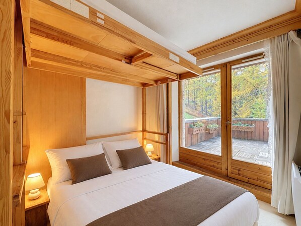 A wooden bedroom with a large bed, a nightstand, and a window with a view of trees. - Charming 4-room apartment for 6 at slopes' edge with wifi (Val-d'Isère)