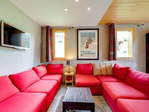 A modern living room with a sectional sofa, a flat-screen TV, a framed poster, and two windows with curtains. - Charming apartment, piste view - 6 guests (Landry)