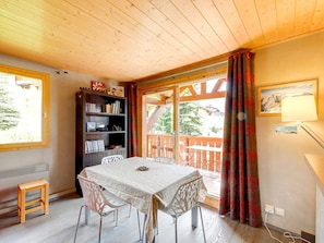 A room with a wooden ceiling, a table with chairs, a bookshelf, and a window with curtains.