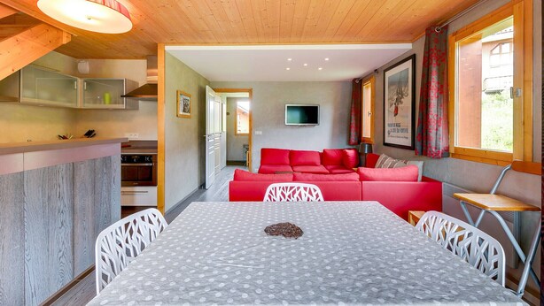 Une pièce chaleureuse avec un plafond en bois, une table à manger recouverte d’une nappe à pois, un canapé rouge et un téléviseur fixé au mur.