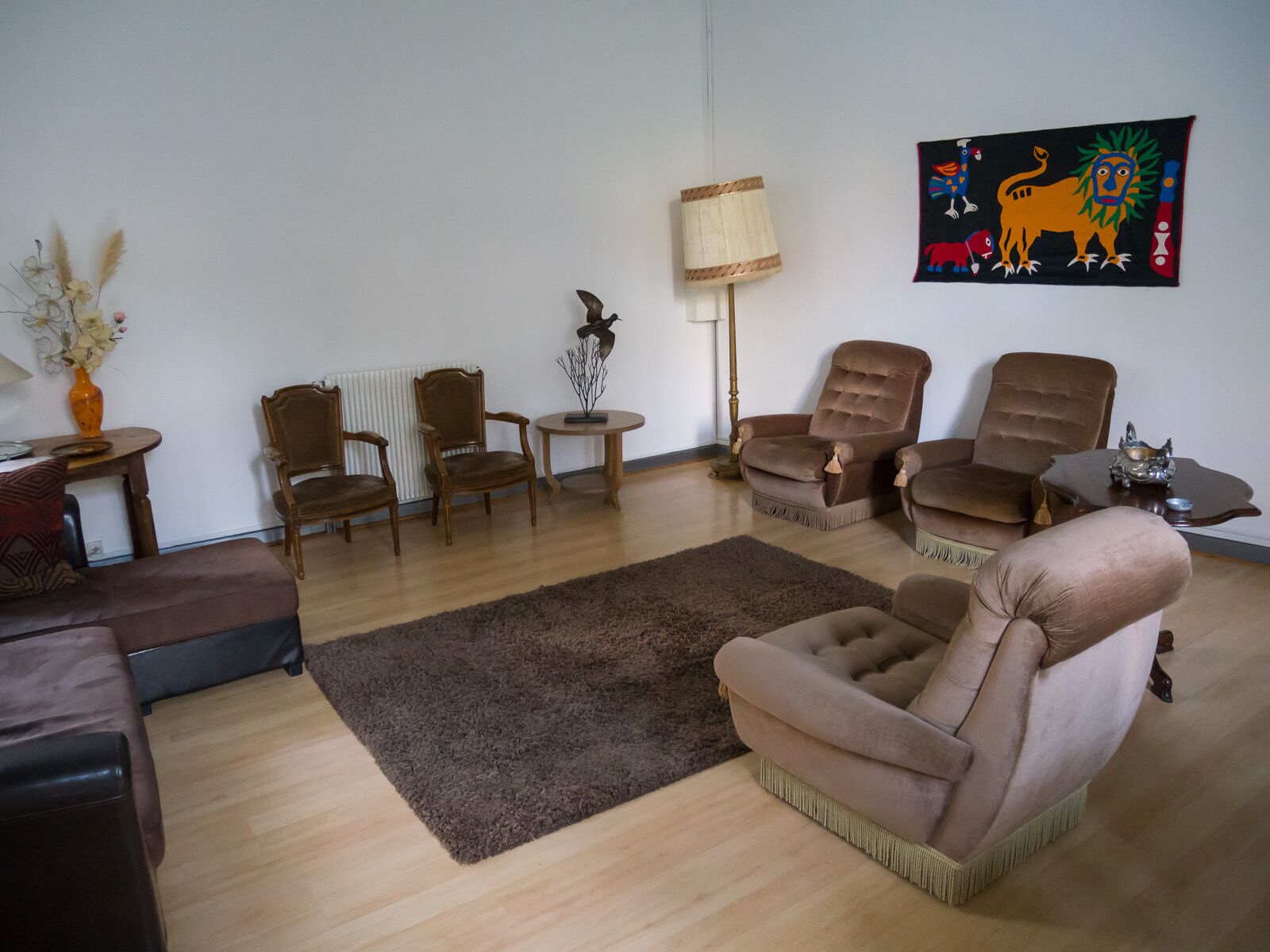 Ein Wohnzimmer mit einem braunen Sofa, zwei Holzstühlen, einem kleinen runden Tisch und einer Stehlampe.