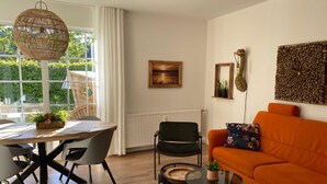 Een moderne woonkamer met een eethoek, met een ronde tafel, stoelen, een oranje bank en een groot raam met gordijnen.