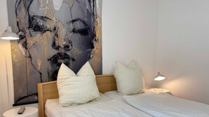 Een net opgemaakt bed met witte beddengoed en twee kussens, een rond nachtkastje met een afstandsbediening, een lamp op het nachtkastje, en een groot abstract schilderij aan de muur.