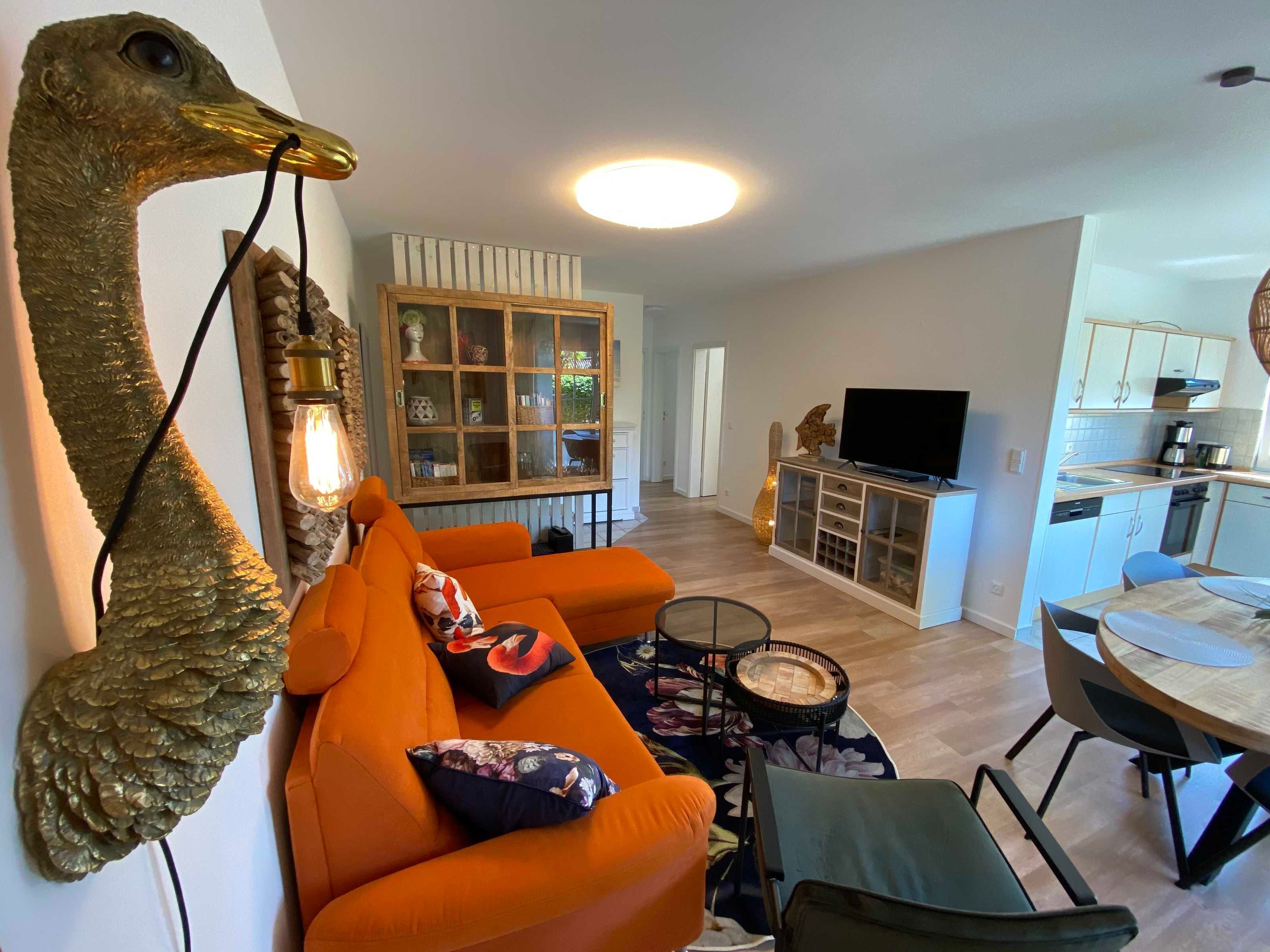 Un salon avec un canapé orange, une grande sculpture d’oiseau décorative, un meuble en bois doté de portes en verre et un coin repas avec une table ronde et des chaises.