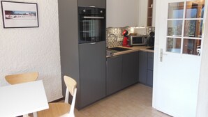 Una cocina compacta con horno integrado, microondas y lavamanos. Hay una pequeña mesa de comedor con sillas y un cuadro en la pared.