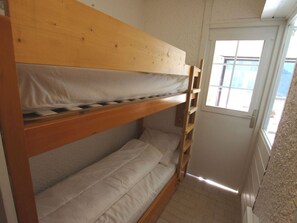 Una cama tipo litera con estructura de madera y ropa de cama blanca en una habitación con suelo de baldosas y una puerta con ventanal.